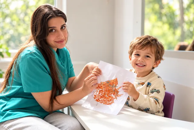 An educator and smiling child show a craft beside a window.